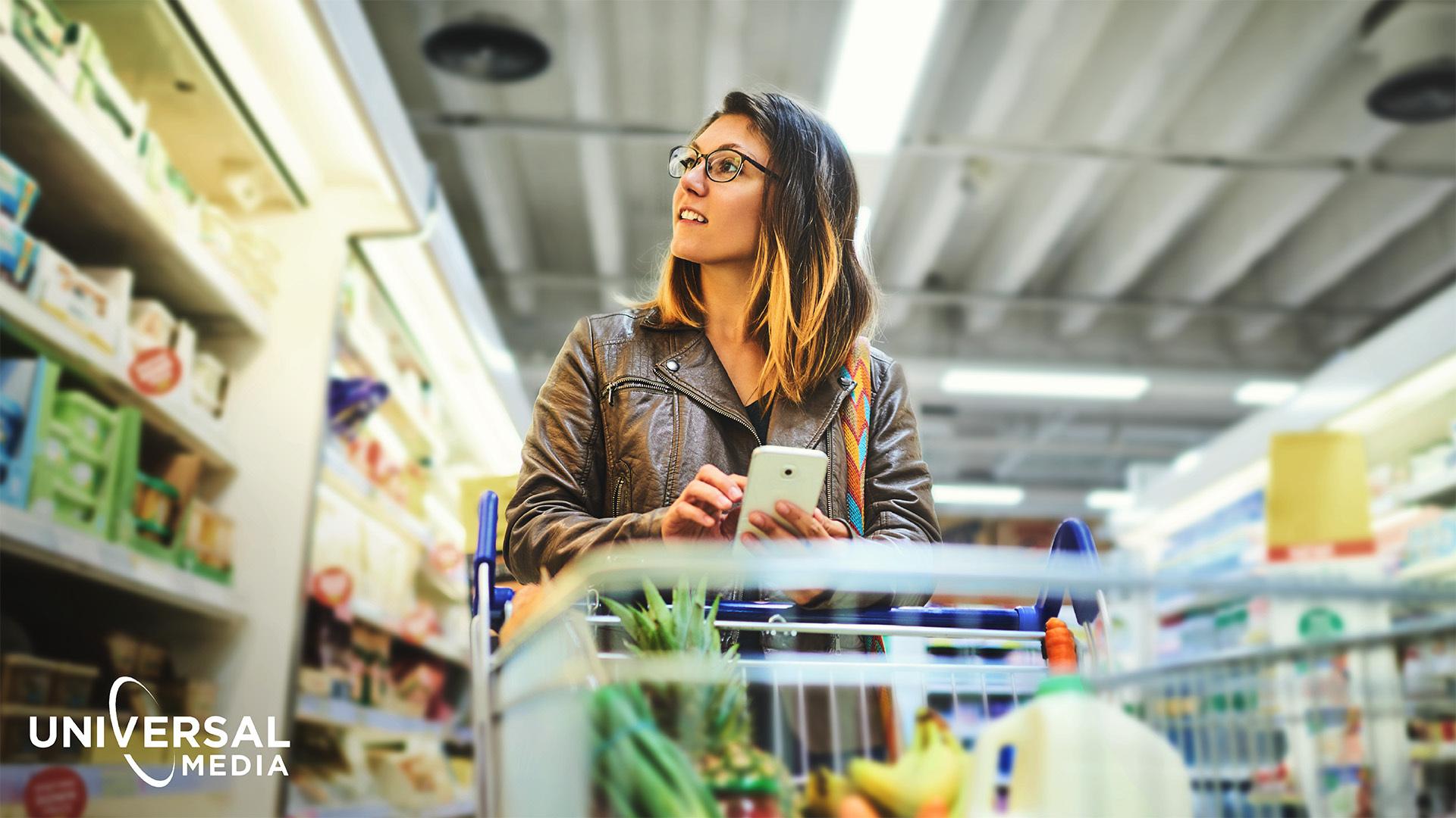 Image of a woman shopping in a grocery store while holding her phone and the "Universal Media" logo in the bottom left corner