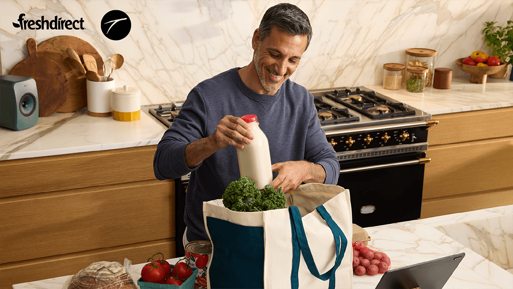 Image of a man pulling groceries out of a tote bag, in his kitchen with the FreshDirect logo locked in the top left corner